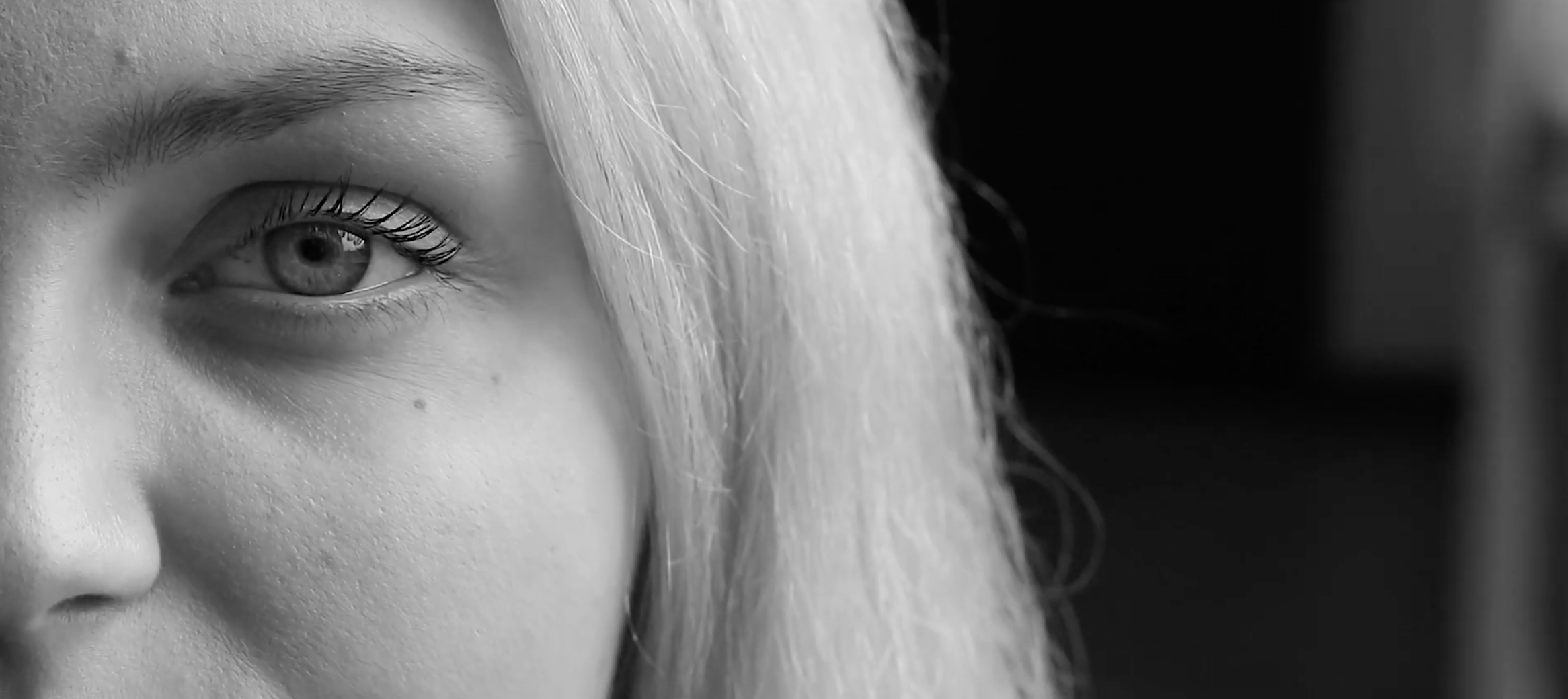 Close-up of a woman's face showing her eye, eyebrow, and part of her nose, with light-colored hair partially covering her face, in black and white.