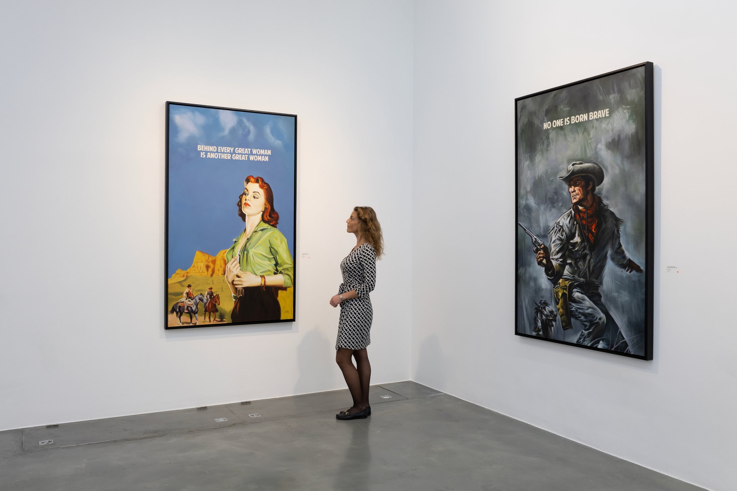 A woman with curly blonde hair in a patterned dress looking at framed posters in an art gallery. The left poster features a stylized woman with red hair and a quote, the right poster shows a man in Western attire with a gun and a quote.
