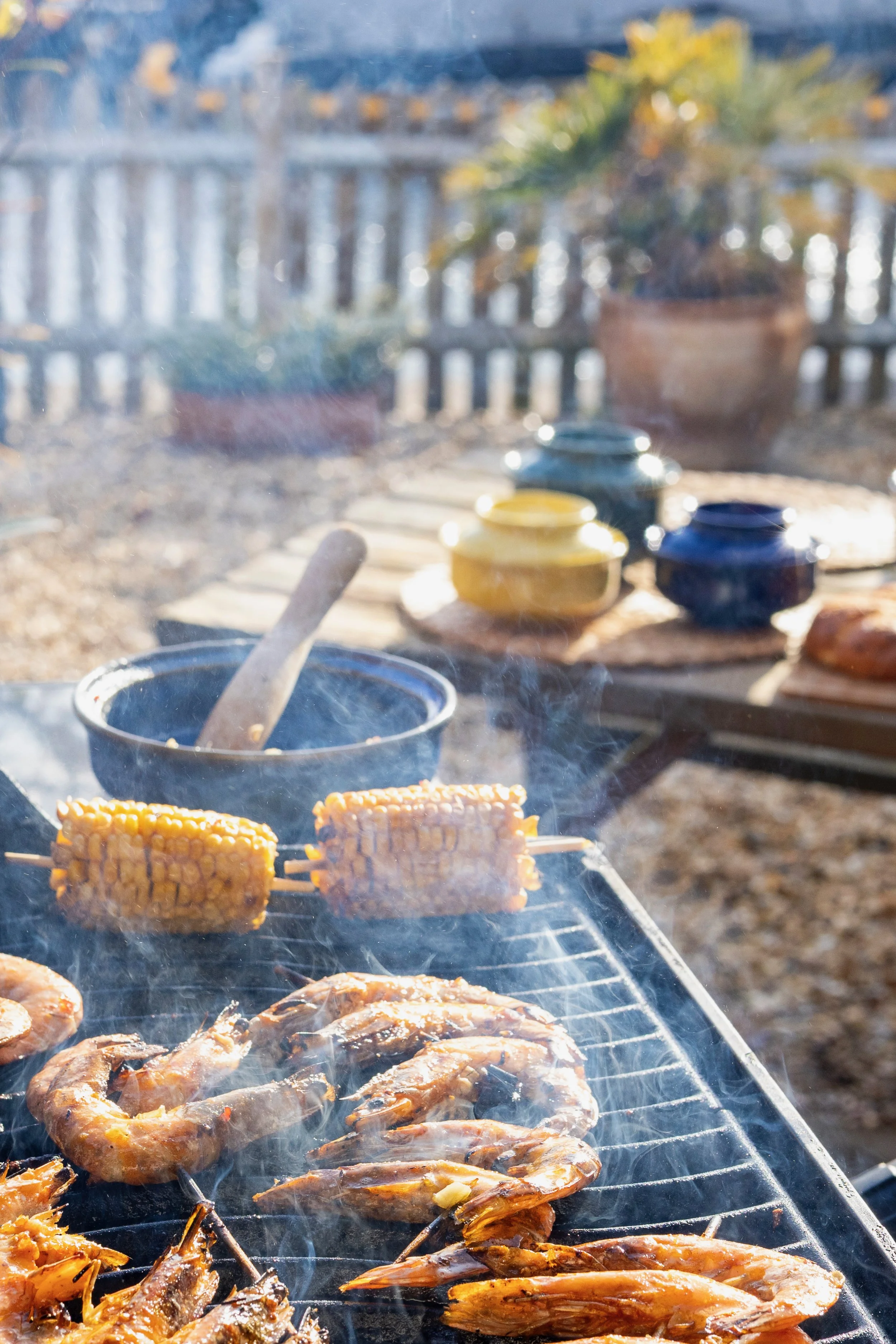 Outdoor scene with grilled shrimp and corn on the cob cooking on a barbecue grill. In the background, there are decorative pots with plants. The image is slightly out of focus.