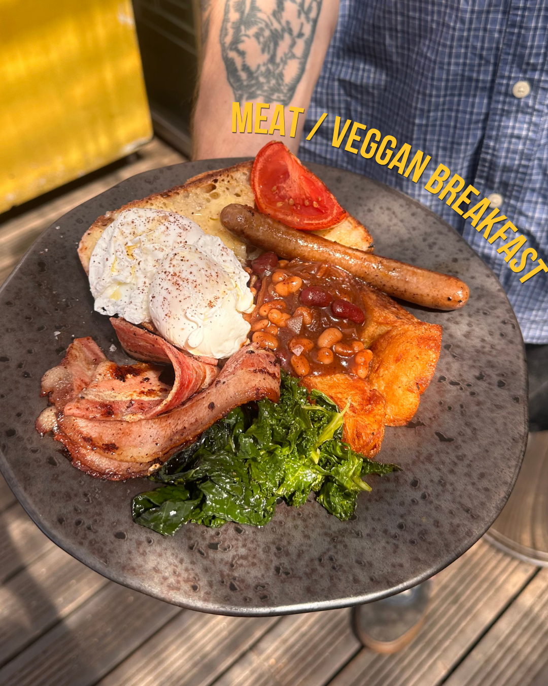 A black plate with vegan breakfast including a poached egg, tomato slice, sausage, baked beans, bacon, and greens held by a person in a blue plaid shirt.