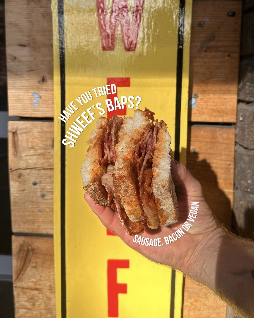 Close-up of a sandwich with sausage, bacon, and possibly vegan ingredients, held in a hand over a yellow and black cardboard sign.