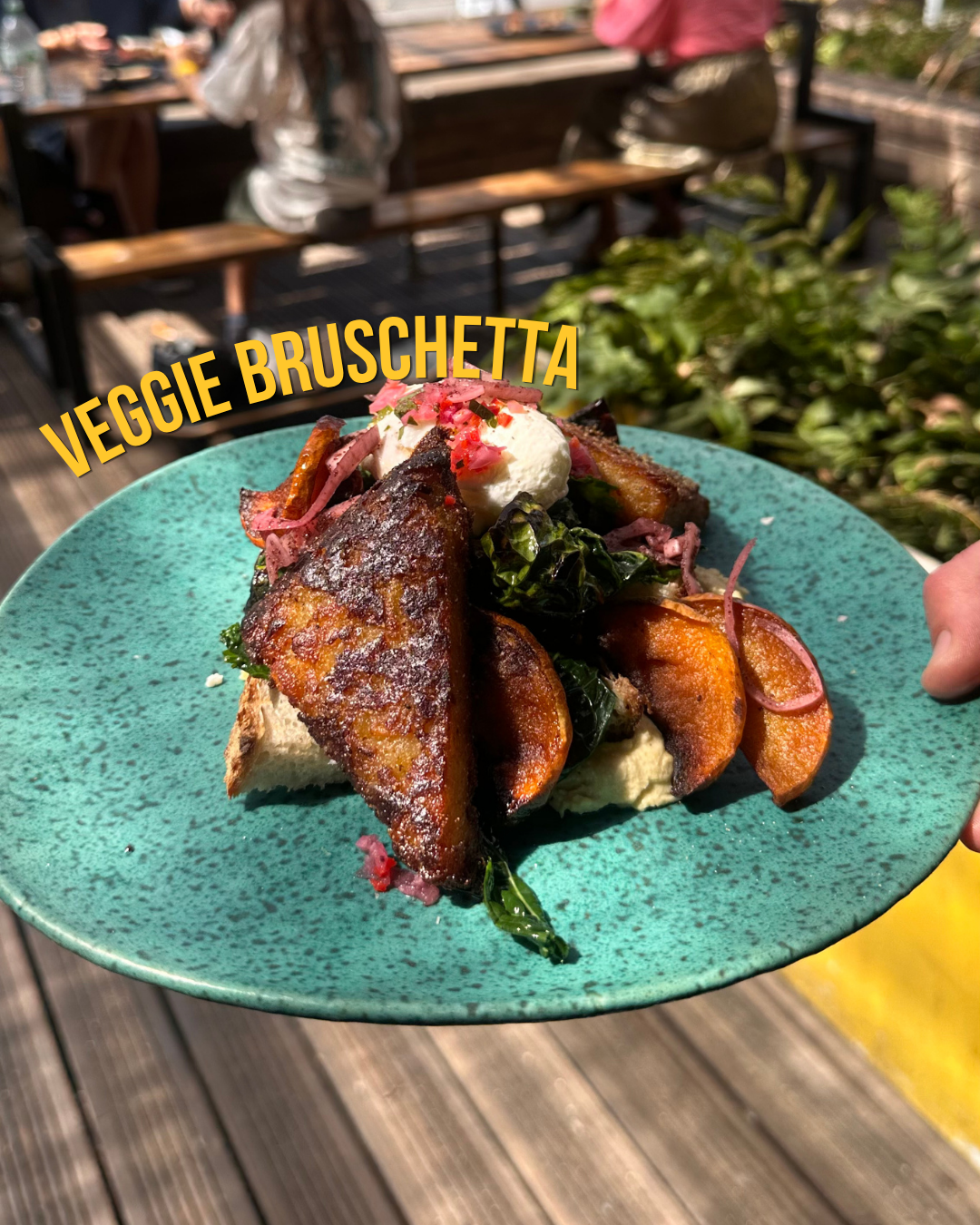 A turquoise plate holds a veggie bruschetta with roasted vegetables, greens, cheese, and pink toppings. The background shows people sitting outdoors, suggesting a patio or outdoor dining area.