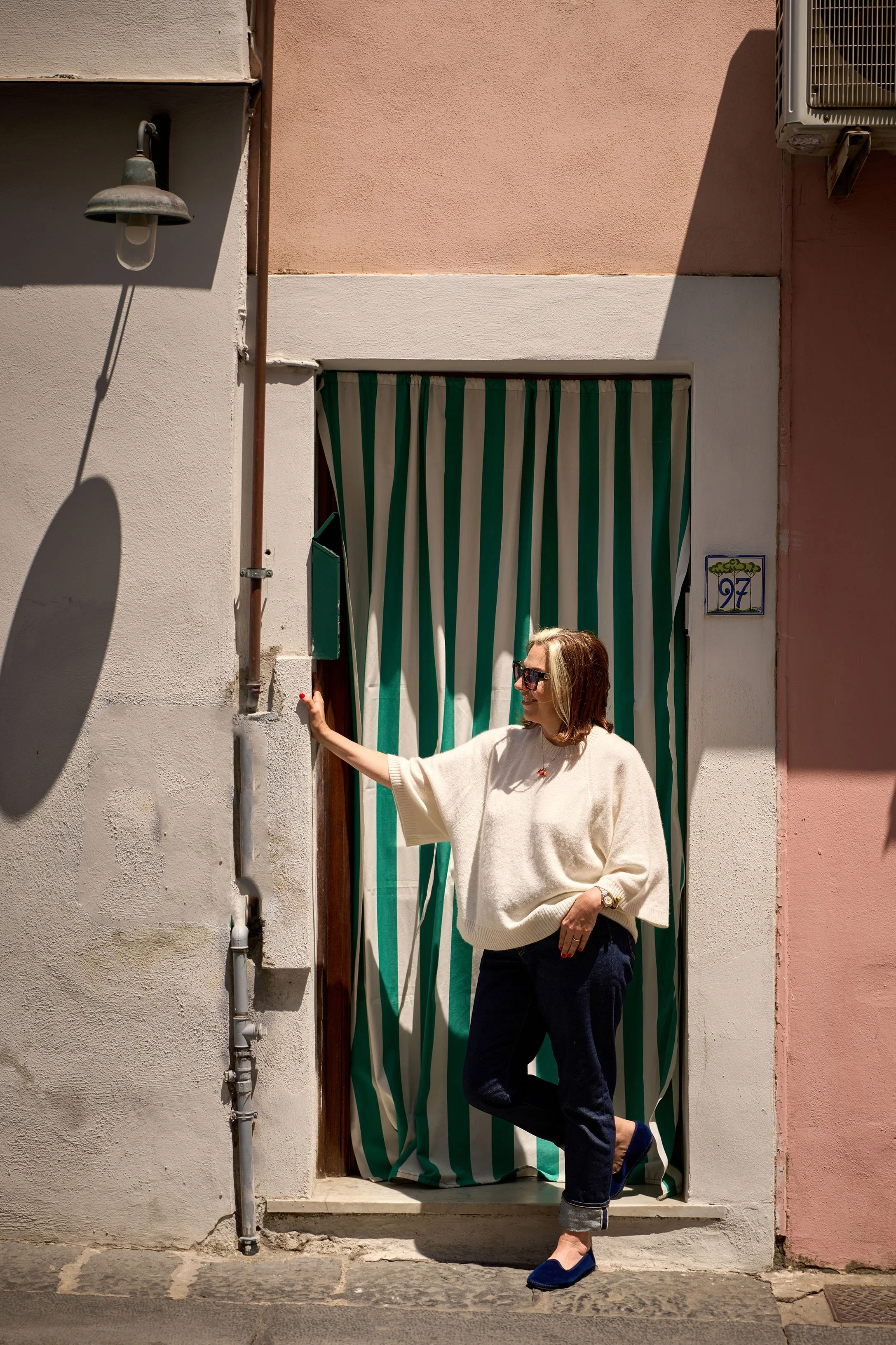 A woman dressed in a beige sweater and dark pants is standing on a stone sidewalk outside a building with a pink and white wall. She is smiling and wearing sunglasses, with her right hand holding a green door handle and her left hand in her pocket. The scene is sunny, with shadows cast on the wall, and there is a striped green and white curtain behind her.