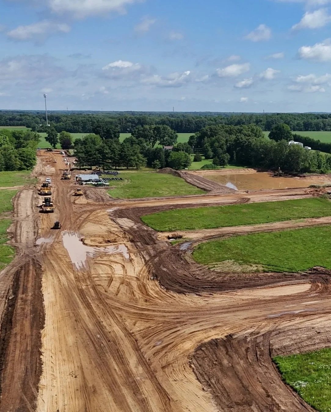 Construction site with dirt roads, construction vehicles, and equipment, surrounded by green fields and trees under a partly cloudy sky.