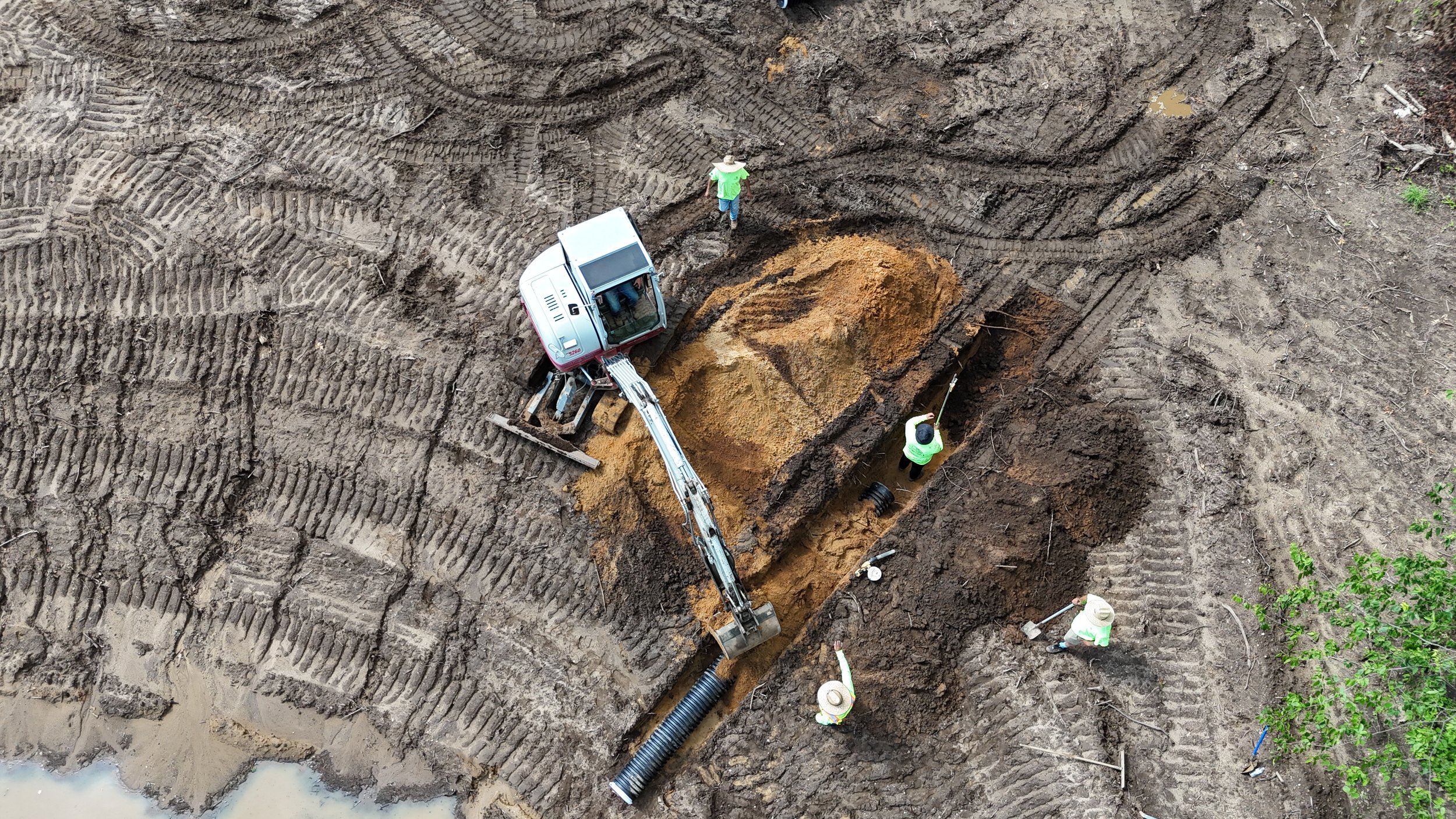 An aerial view of a construction site with workers operating machinery and digging a trench in muddy ground, tire tracks visible on the soil.