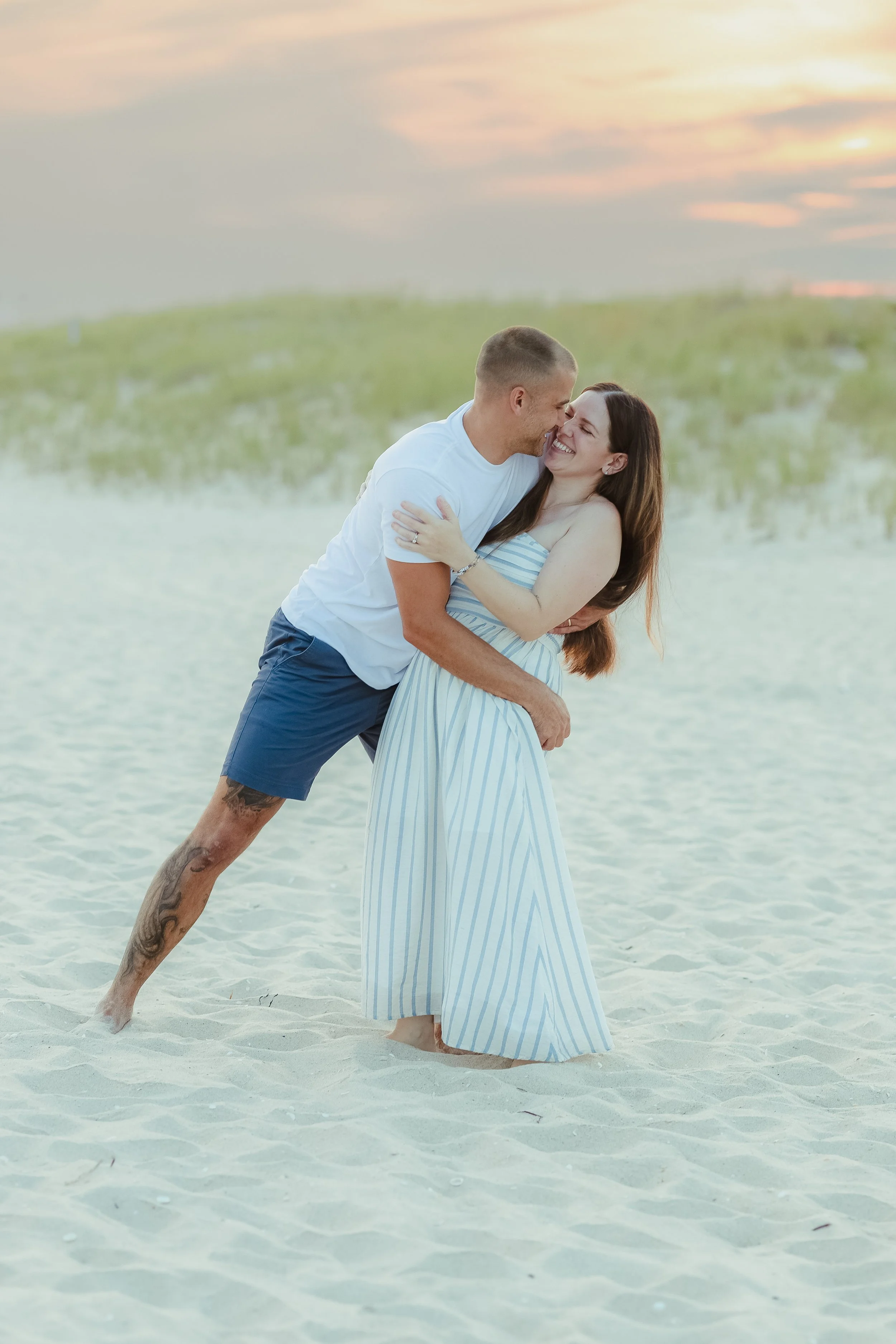A happy couple embracing on a sandy beach at sunset, with the man leaning in while holding the woman and both smiling.