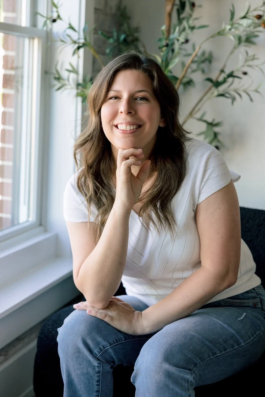 A woman with wavy brown hair sits on a dark couch near a window with white blinds, smiling, wearing a white shirt and blue jeans, with a large leafy plant in the background.