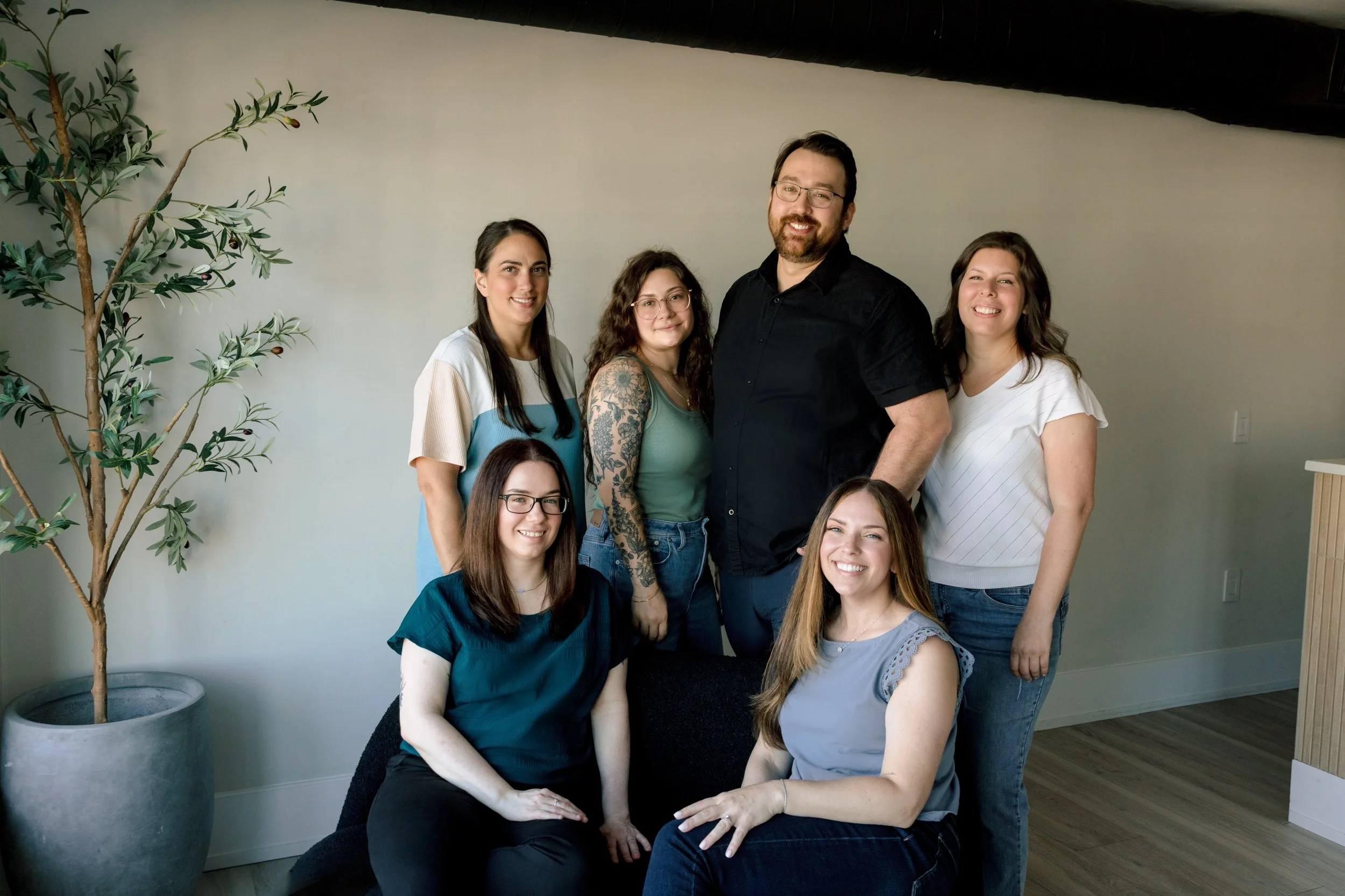Group of seven people, five women and two men, smiling and posing for a group photo in a room with a white wall and a potted tree on the left.