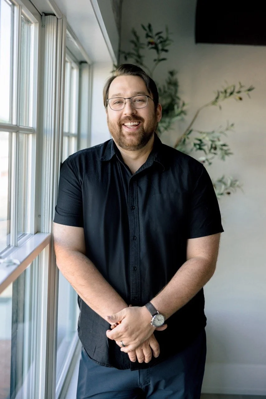 A man with glasses and a beard, smiling, standing indoors near a window with natural light, wearing a black short-sleeve shirt and a watch.