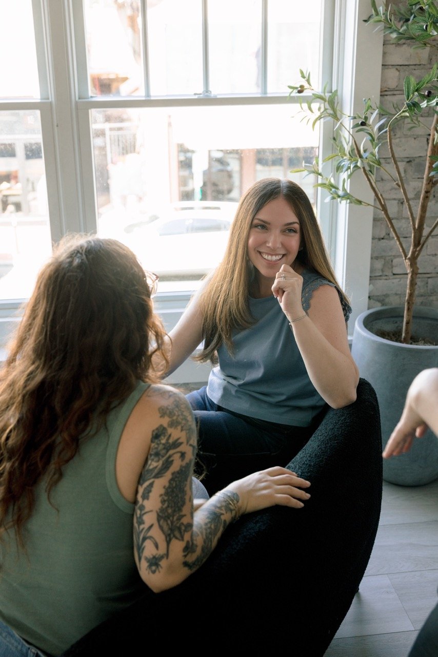 Two women sitting by a large window, engaged in conversation. One woman has light brown hair, is smiling, and wearing a blue top, while the other has curly hair, tattoos on her arm, and is wearing a green tank top.