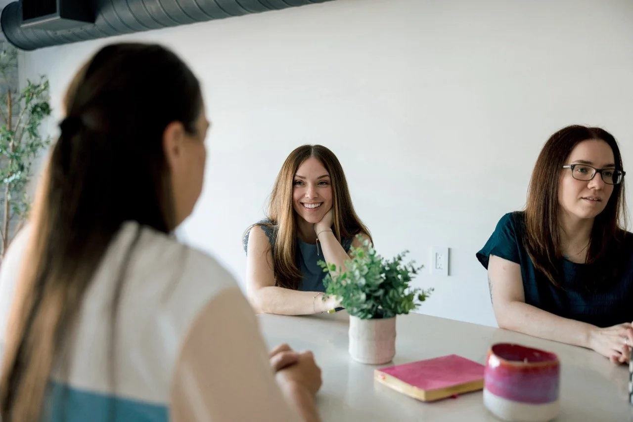 Three women sitting at a table in a casual office setting, with one smiling at the camera and the others engaged in conversation, with a potted plant and notebooks on the table.