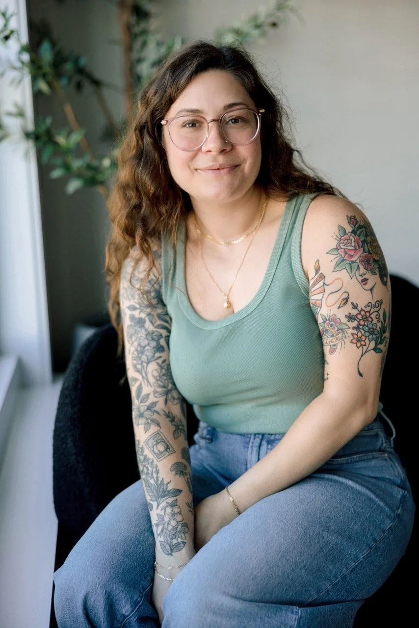 A woman with wavy brown hair, glasses, and tattoos on her arms, sitting indoors near a window with greenery in the background, wearing a green sleeveless top and jeans, smiling softly.