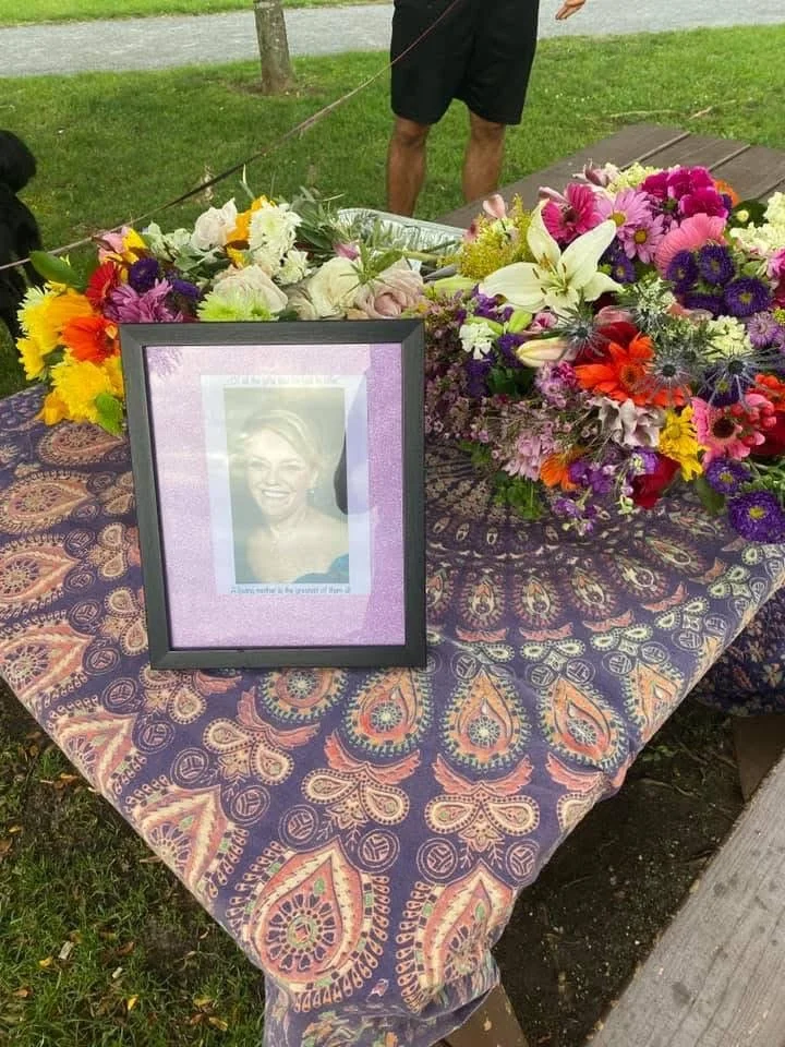 A memorial table with a framed photo of a smiling woman and a colorful flower arrangement on a patterned tablecloth outdoors.