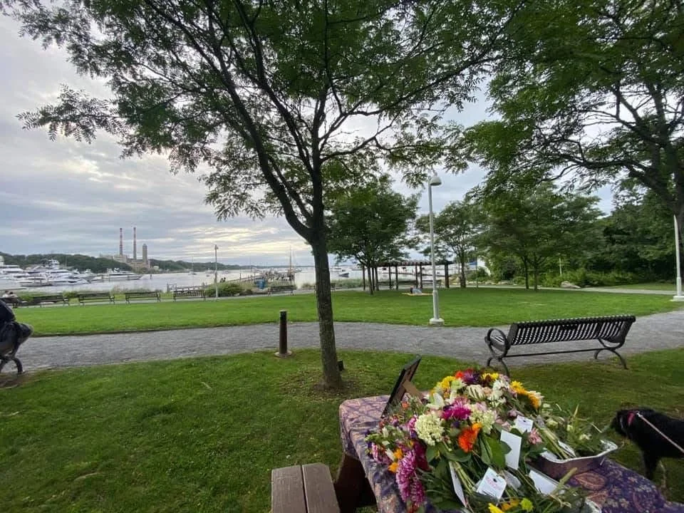 A park with a view of a marina filled with boats, trees, benches, a table with flowers and donation cards, on a cloudy day.