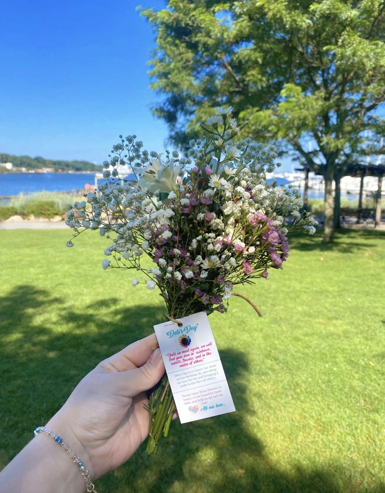 A hand holding a bouquet of small pink and white flowers with a note card attached. In the background, there is a lush green park with trees, a body of water, and a blue sky.