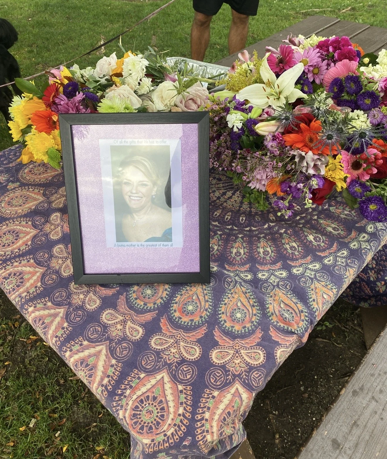 A table covered with a colorful paisley-patterned cloth displaying a framed photograph of a smiling woman and a large arrangement of vibrant flowers outdoors.