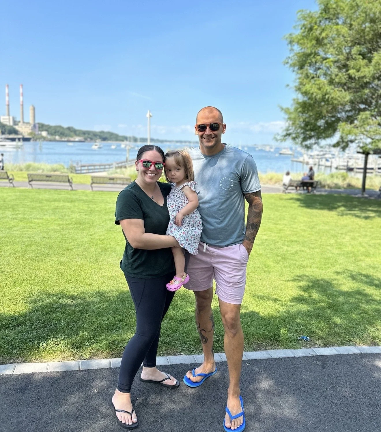 A family of three, a woman, a man, and a young girl, standing together outdoors near the water on a sunny day. The woman is holding the girl, and both are smiling. The man is standing next to them, also smiling, wearing sunglasses and flip-flops. In 