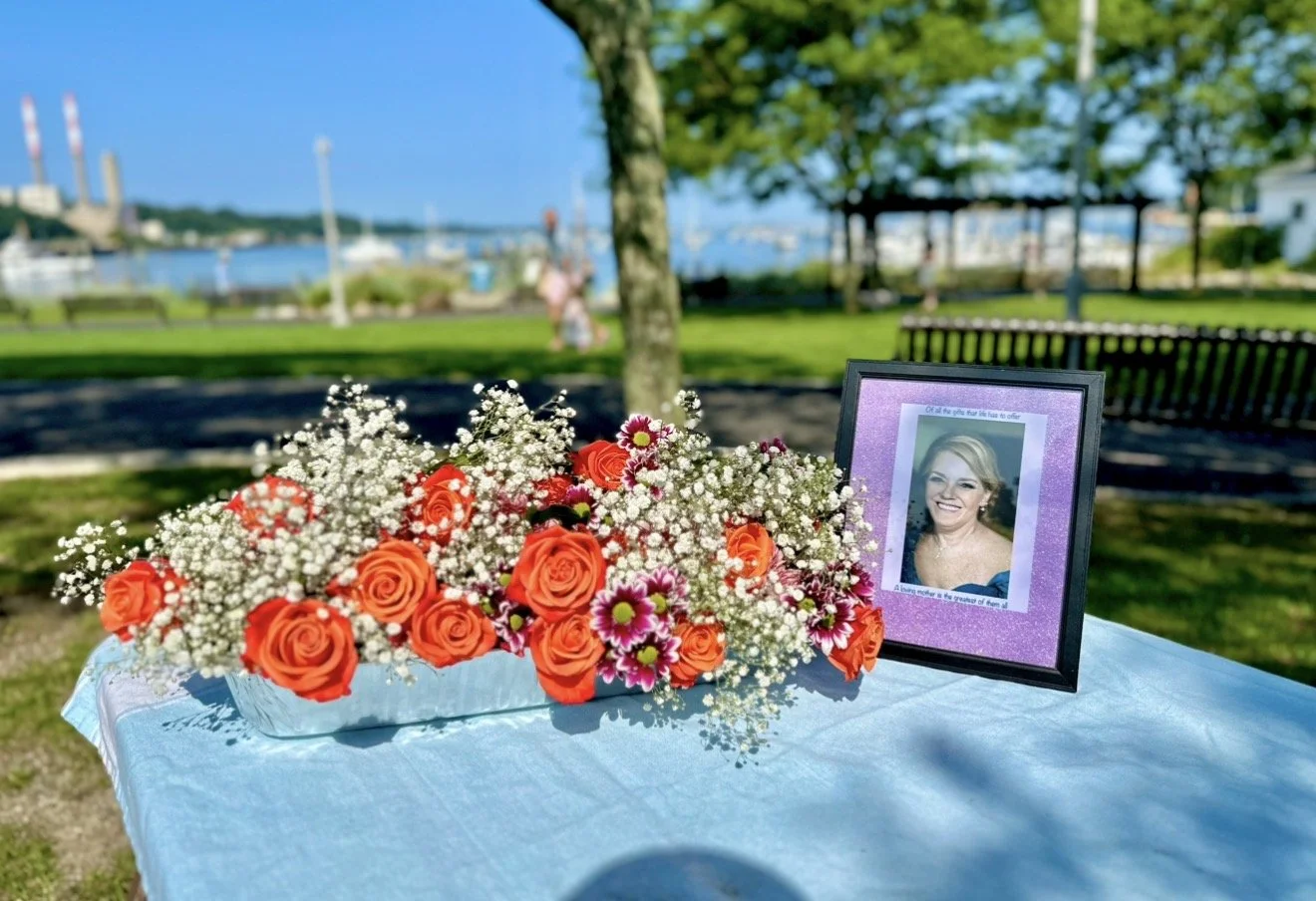 A memorial table with a framed photo of a smiling woman, surrounded by orange roses, pink flowers, and baby's breath, set outdoors near a waterfront park with trees, benches, and boats in the background.
