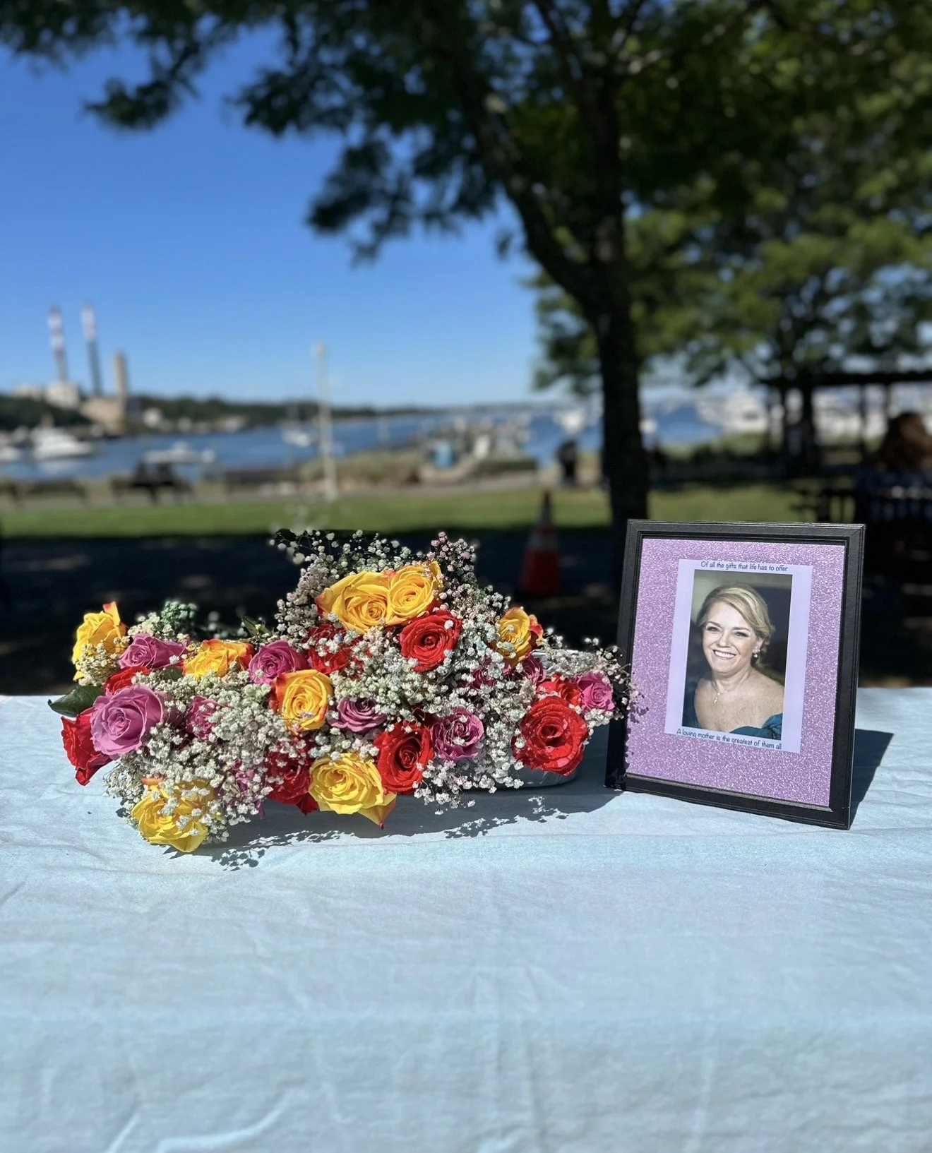 A memorial setup with a bouquet of multicolored roses and a framed photograph of a smiling woman, outdoors by a waterfront with boats and industrial chimneys in the background.