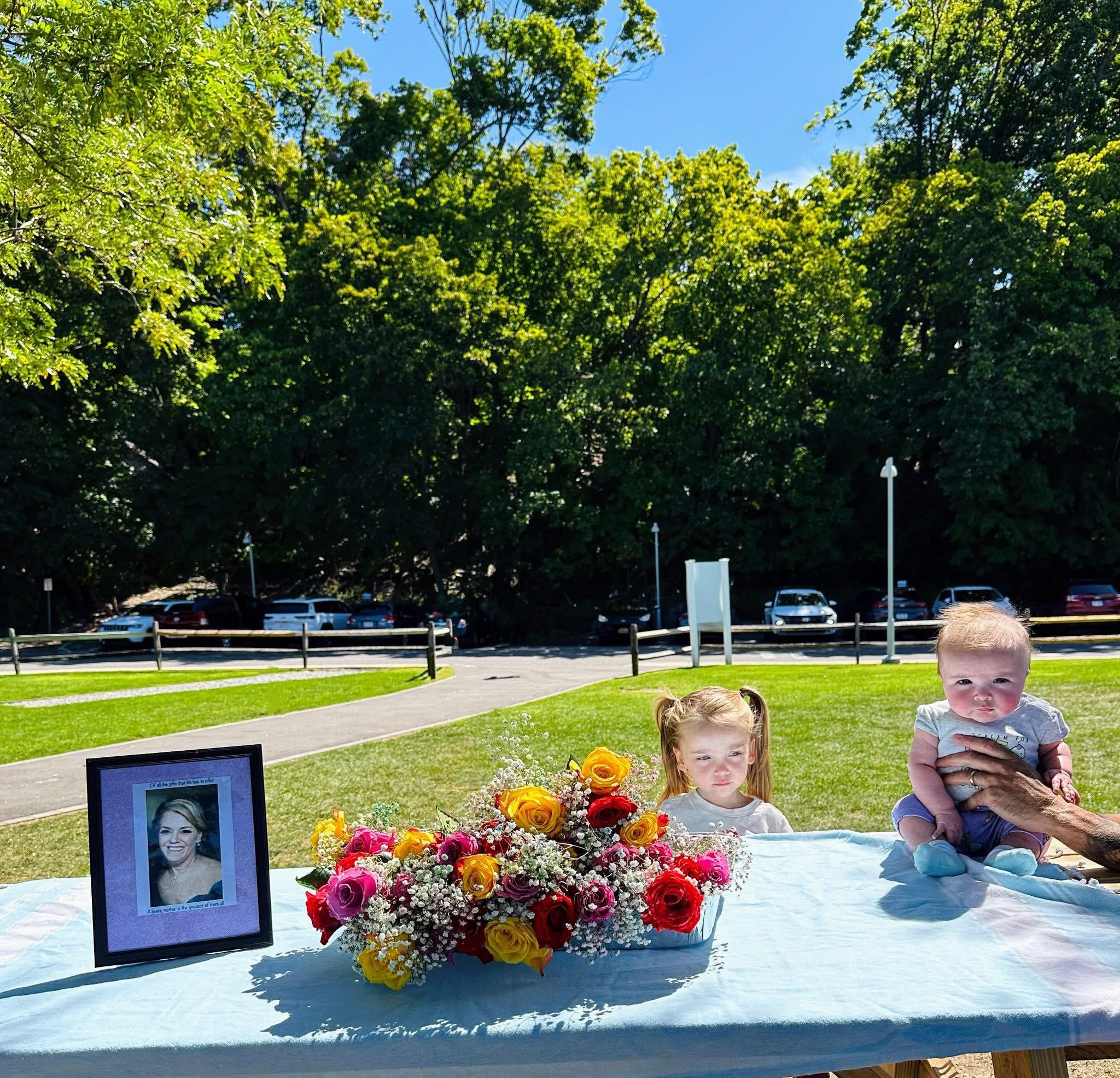 Memorial display with a framed photograph of a woman, surrounded by a large bouquet of colorful flowers, including yellow, pink, and white roses. There are two young children, a girl and a boy, sitting near the display on a table covered with a light
