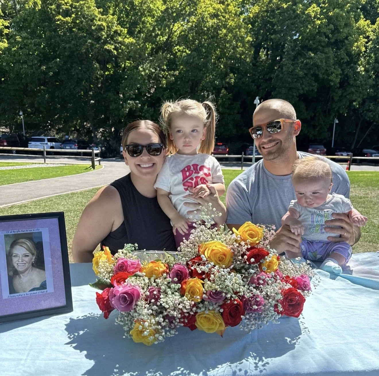 A family of four sitting outdoors at a table with a large bouquet of colorful flowers, smiling and posing for a photo. The woman and man are wearing sunglasses, and two young children are in their laps, one girl with pigtails and one boy. There is a framed photo on the table,.