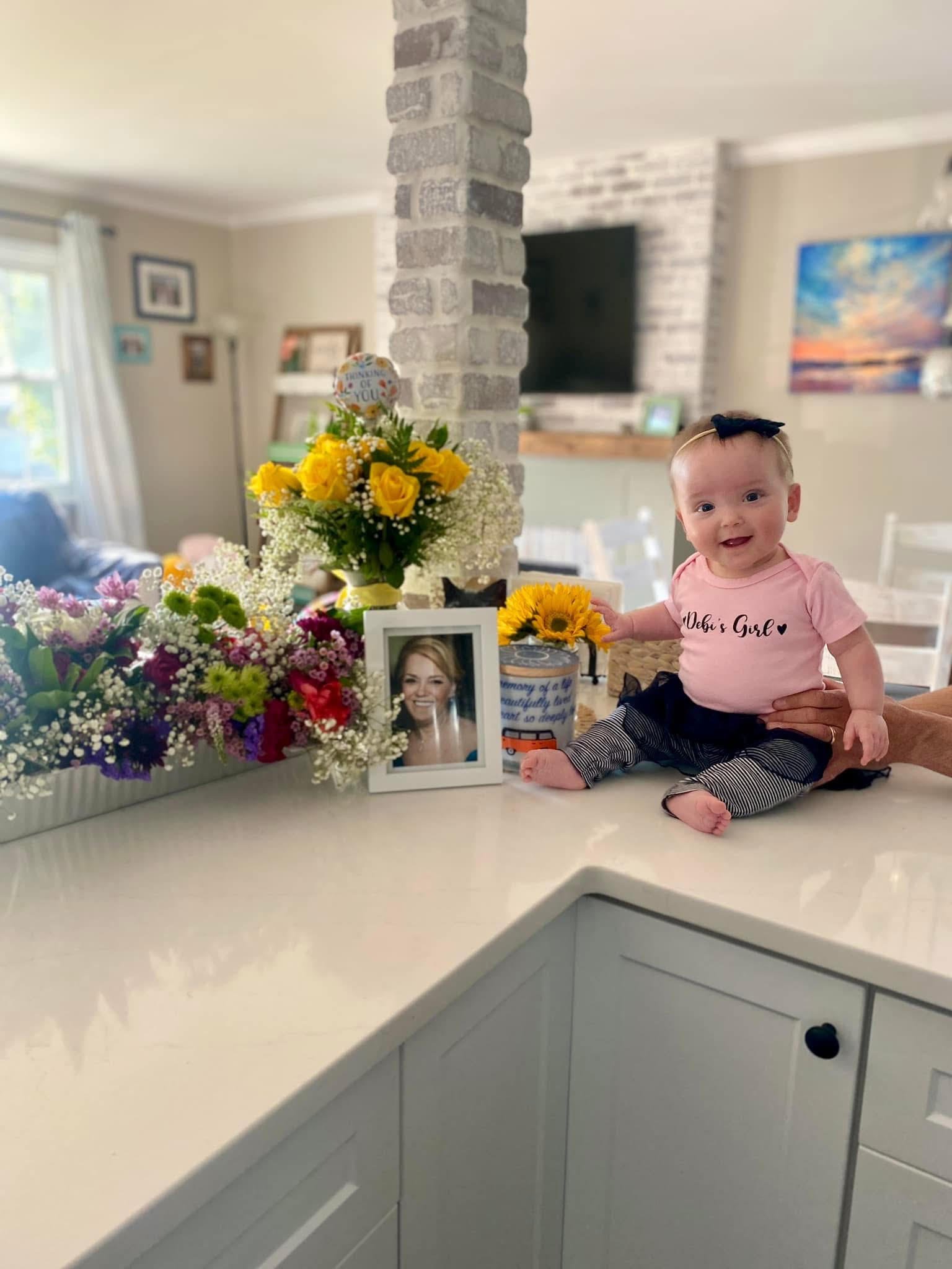 A baby girl with a black bow headband, sitting on a kitchen counter, wearing a pink shirt that says 'Debi's Girl' and striped pants, surrounded by colorful flowers and a framed photo at a celebration or memorial with a bouquet and a card.