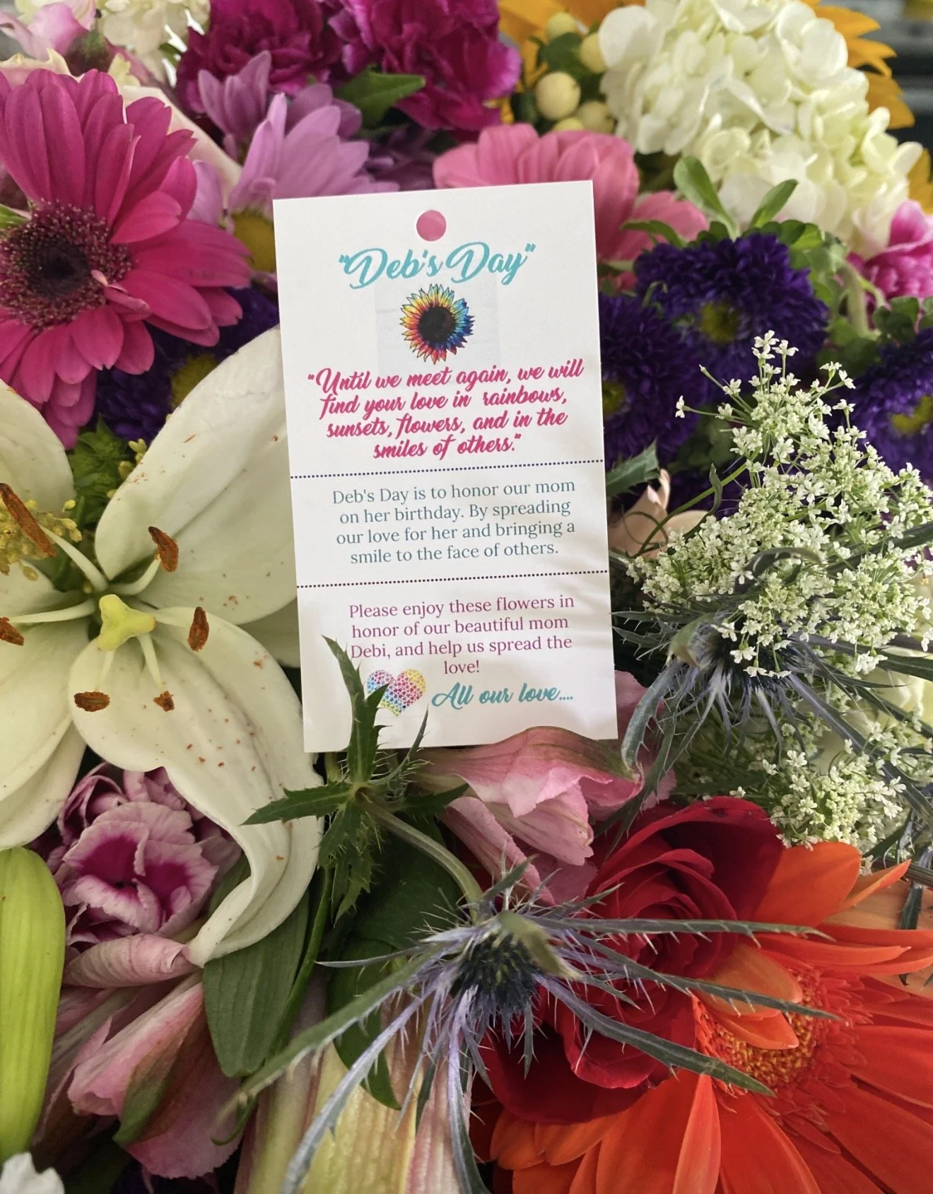 A bouquet of various colorful flowers including white lilies, pink roses, purple and white filler flowers, and orange blooms. A card is placed among the flowers with a message for Deb's Day, honoring their mom.