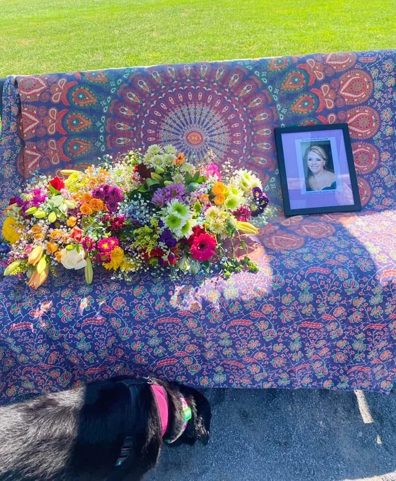 Memorial setup with colorful flowers, a framed photo of a smiling woman, and a decorative tablecloth on an outdoor table.
