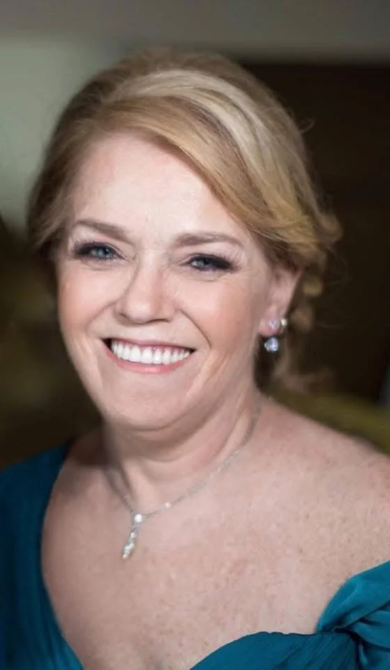 Close-up of a smiling woman with blonde hair, wearing earrings and a necklace, dressed in a teal top.