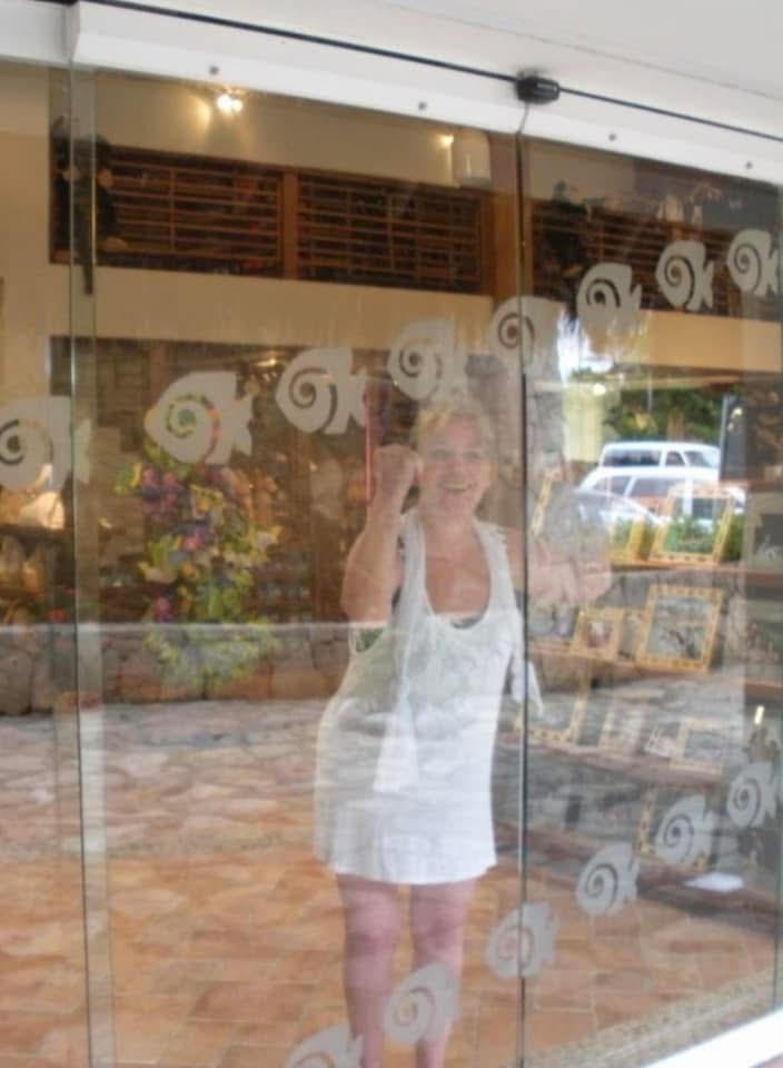 A woman in a white sleeveless dress is smiling and talking on a mobile phone behind a glass door on which decorative spiral patterns are applied. She appears to be indoors, with a flower arrangement and magazine racks visible in the background.