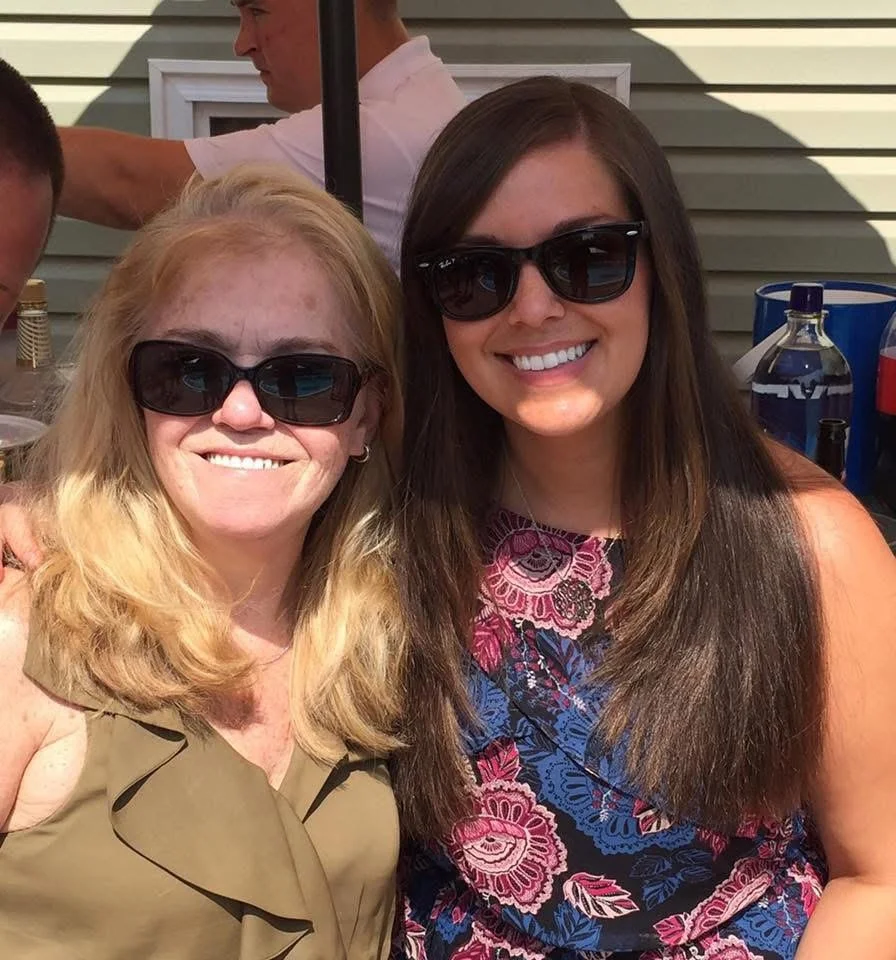 Two women smiling and wearing sunglasses, sitting outside at a gathering or party.