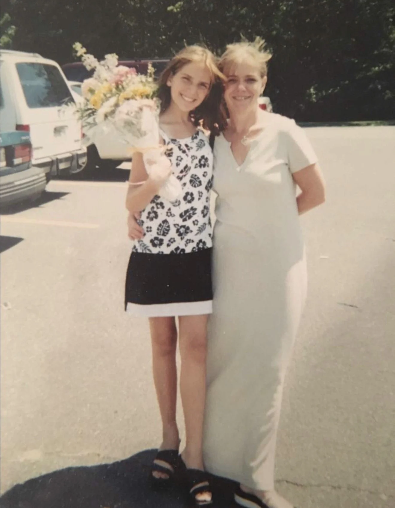 A young woman holding a large bouquet of flowers posing with an older woman in an outdoor parking lot.
