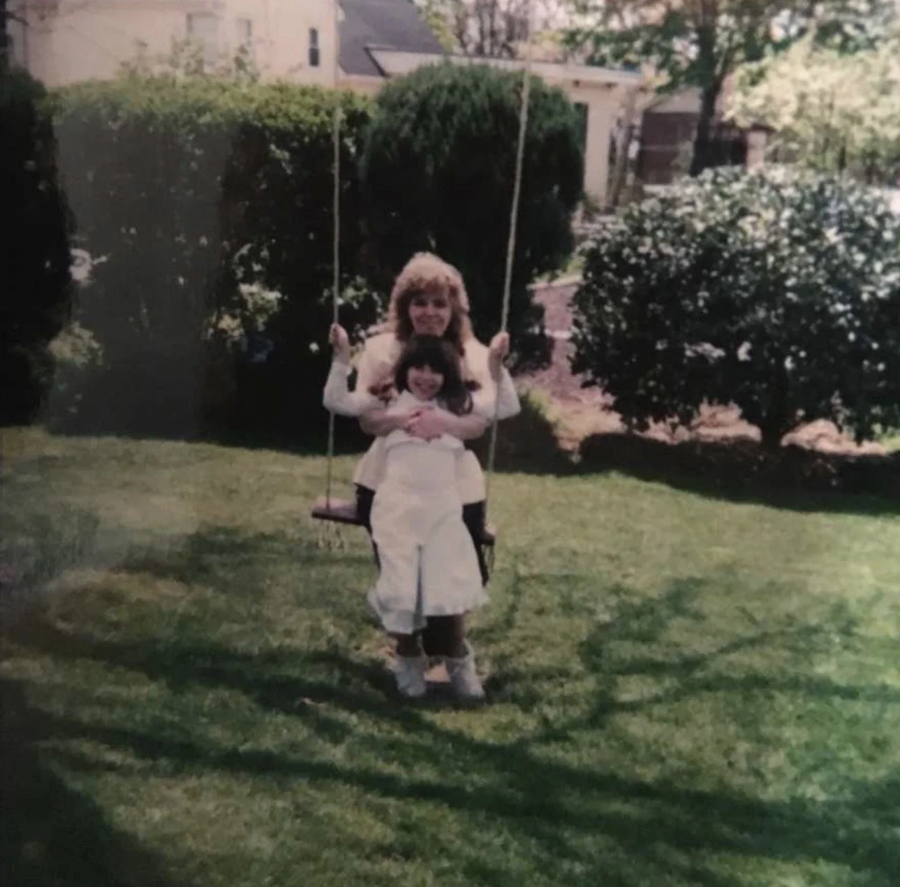 A woman and a young girl on a swing set in a backyard garden with green grass, bushes, and trees.