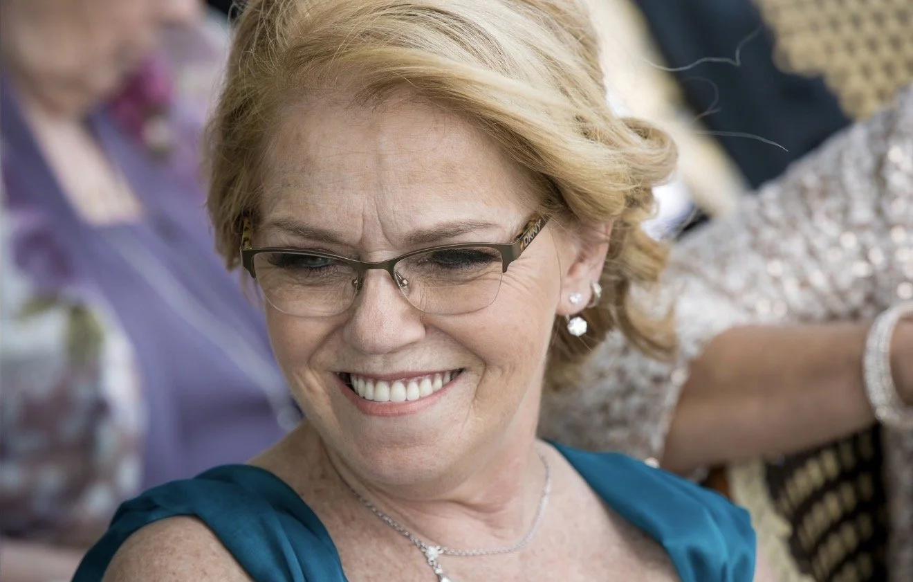 Close-up of a smiling woman with glasses, blonde hair styled in curls, wearing earrings, a necklace, and a teal dress, at what appears to be a social gathering.