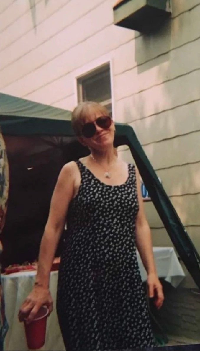 A woman wearing sunglasses and a black and white polka dot dress holding a red cup, standing outdoors near a building with a white wall.