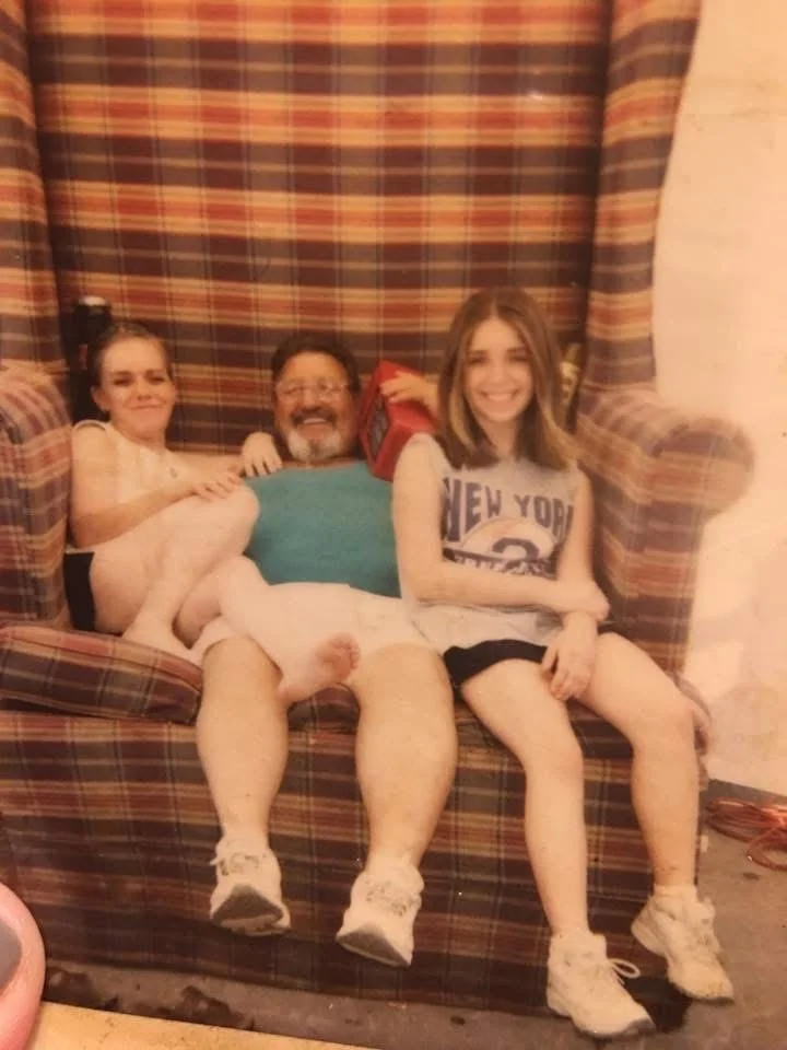 A man with two young girls sitting on a plaid couch with a striped curtain behind them, all smiling and appearing happy.