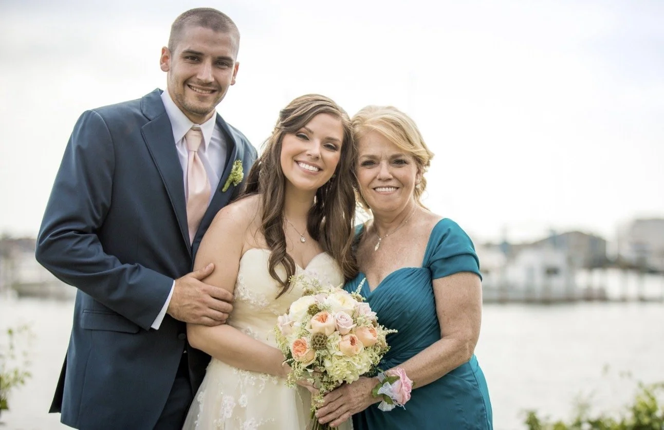 A wedding photo of a bride in a white gown, a groom in a blue suit, and an older woman in a blue dress holding a bouquet of flowers, standing outdoors near a body of water.