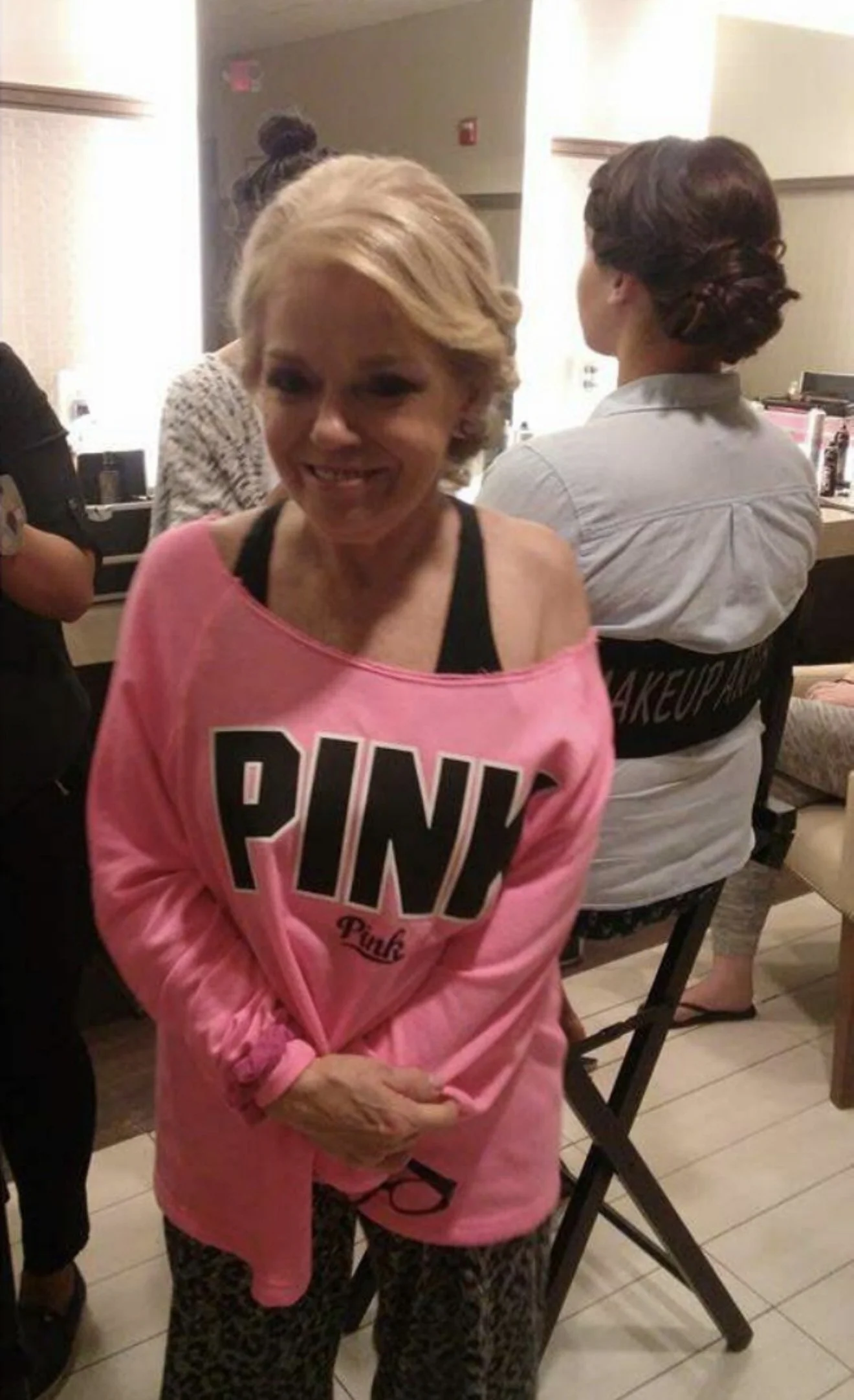 A woman with blonde hair in a short, wavy bob wearing a pink sweatshirt with 'PINK' written on it, standing indoors with her hands clasped in front of her. Behind her are a woman with dark hair tied back and a person with hair in a bun, both facing a