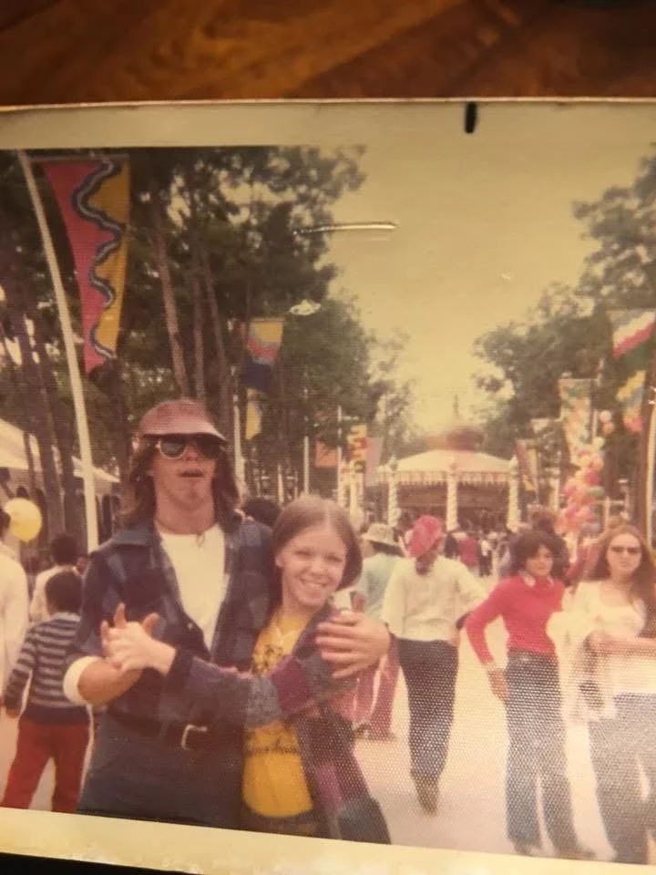 Vintage photo of two women at an outdoor event, standing close together and smiling, with flags and people walking in the background, possibly at a carnival or festival.