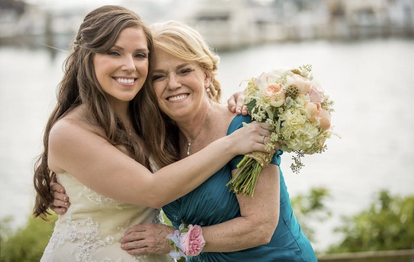 A young woman in a wedding dress and an older woman in a teal dress hugging and smiling outdoors near a body of water, with the younger woman holding a bouquet of flowers.