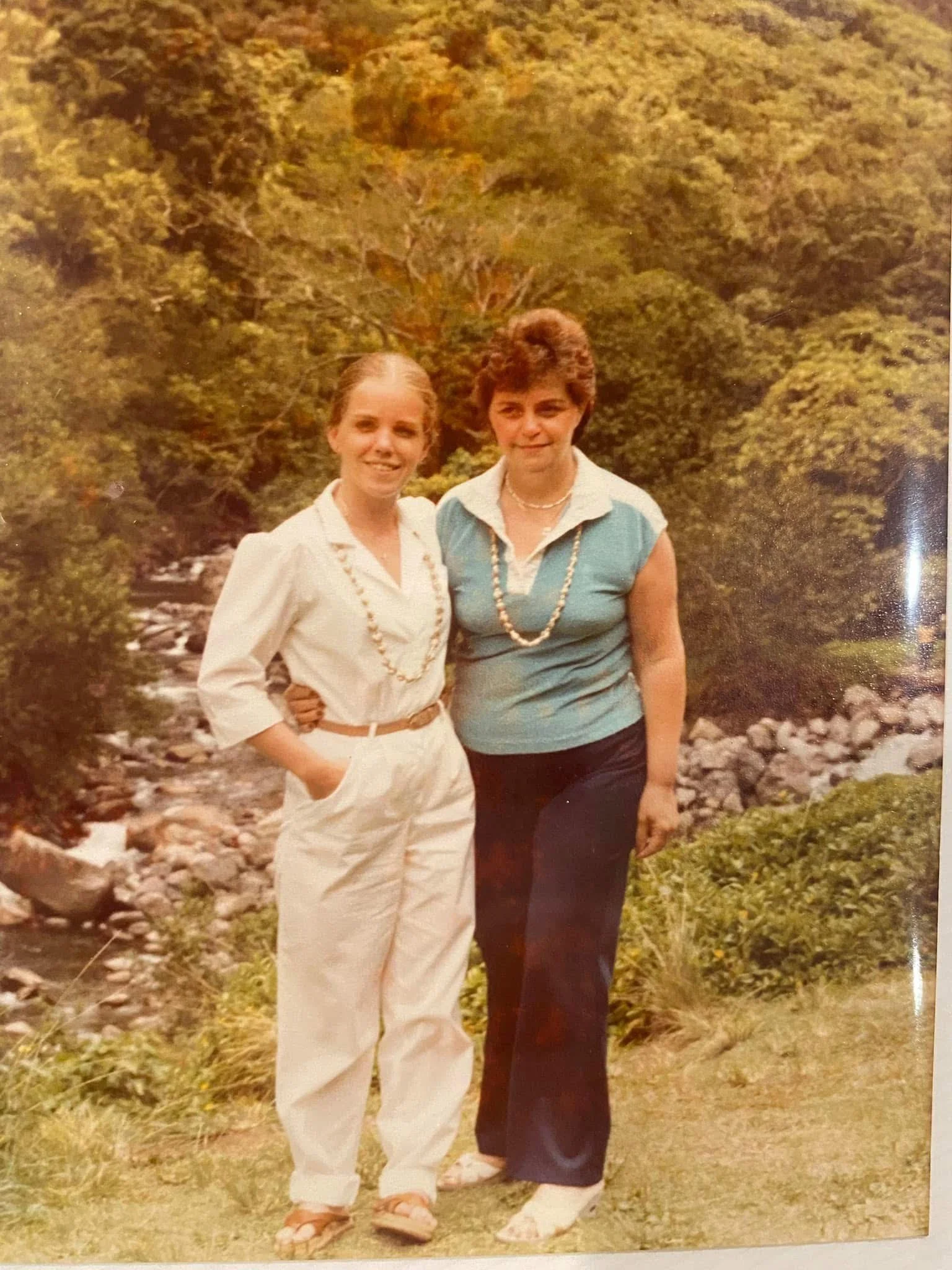 Two women standing outdoors on a rocky and grassy area with greenery and trees behind them, smiling at the camera.