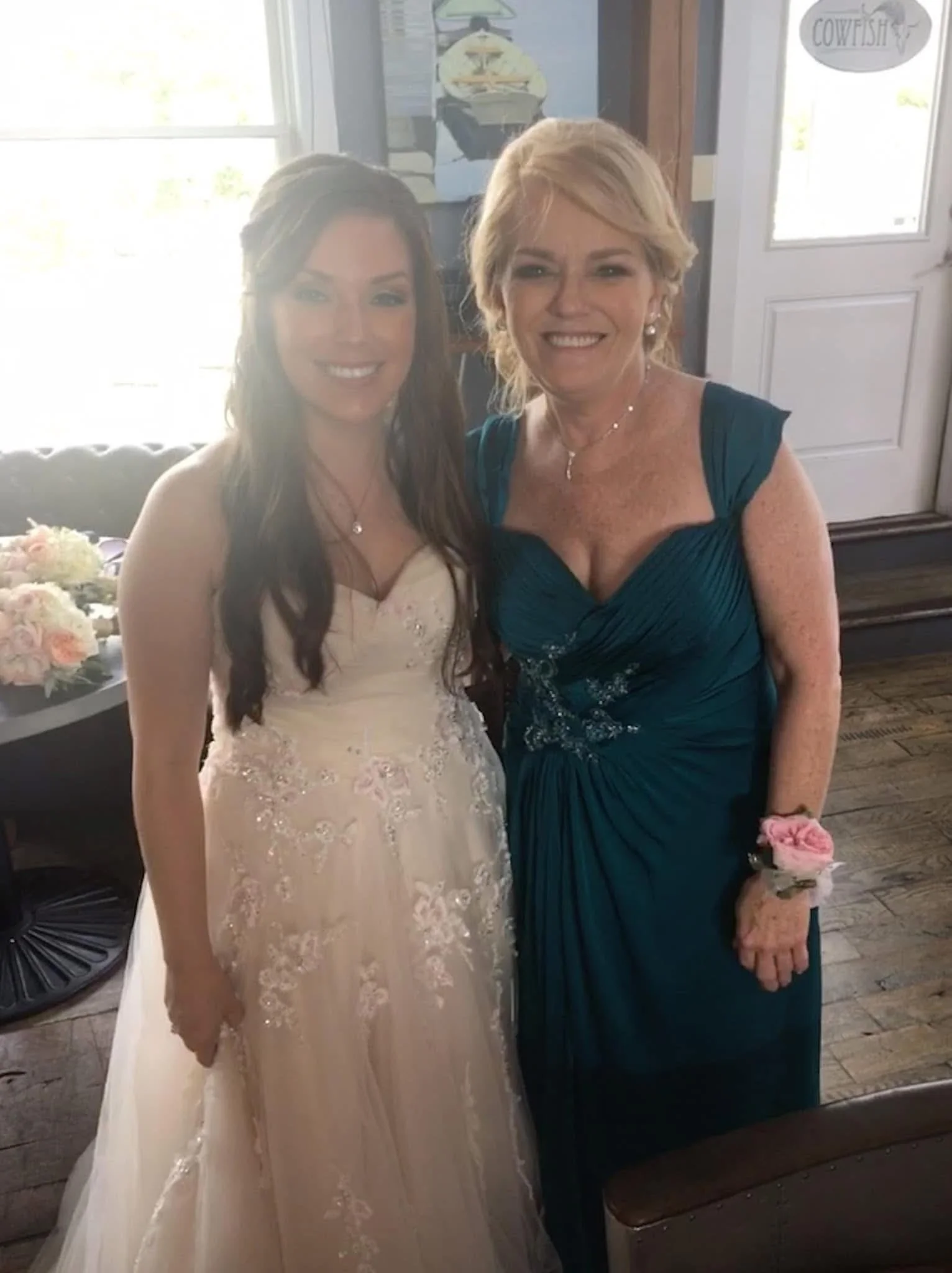 Two women smiling at a wedding reception, one in a white wedding gown and the other in a teal dress, standing indoors near a window with a sign that says "Cowgirl" visible on the door behind them.