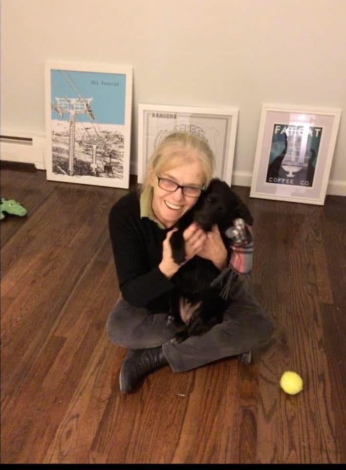 An elderly woman with glasses sitting on a wooden floor, happily hugging a black dog. Behind her are framed pictures leaning against the wall. A yellow tennis ball is on the floor nearby.