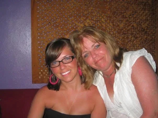 Two women sitting close together and smiling, with a purple wall and orange patterned background behind them.