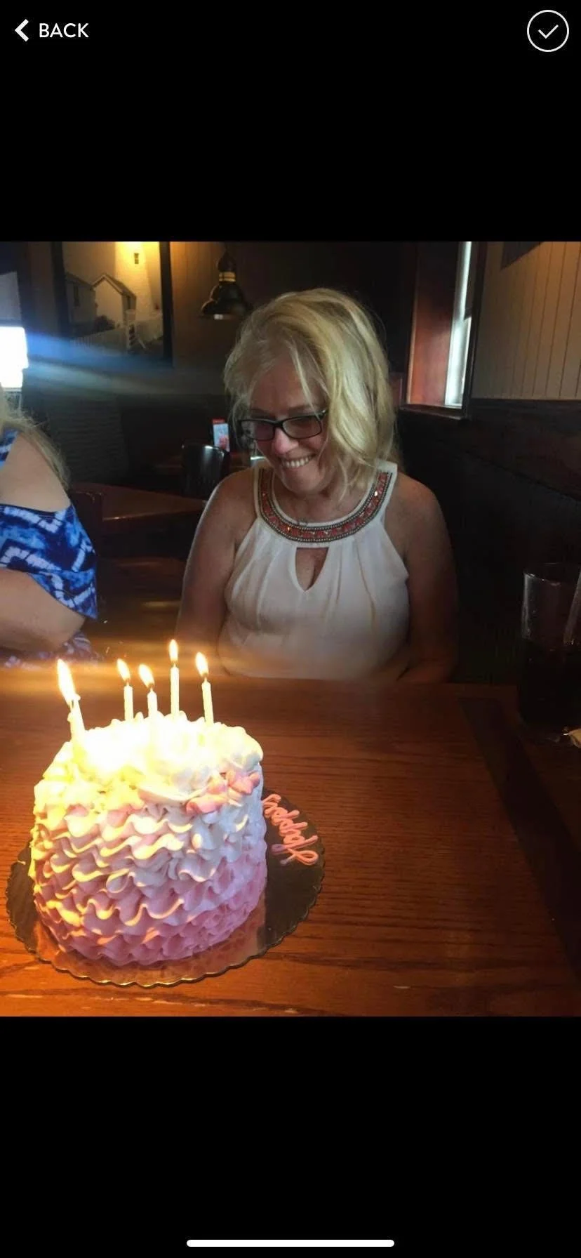 A woman with blonde hair and glasses smiling at a birthday cake with lit candles in a restaurant setting.
