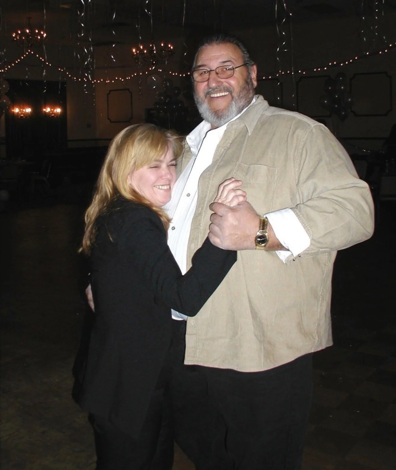 A man and a woman dancing together, smiling, in a dimly lit room decorated with Christmas lights and balloons.