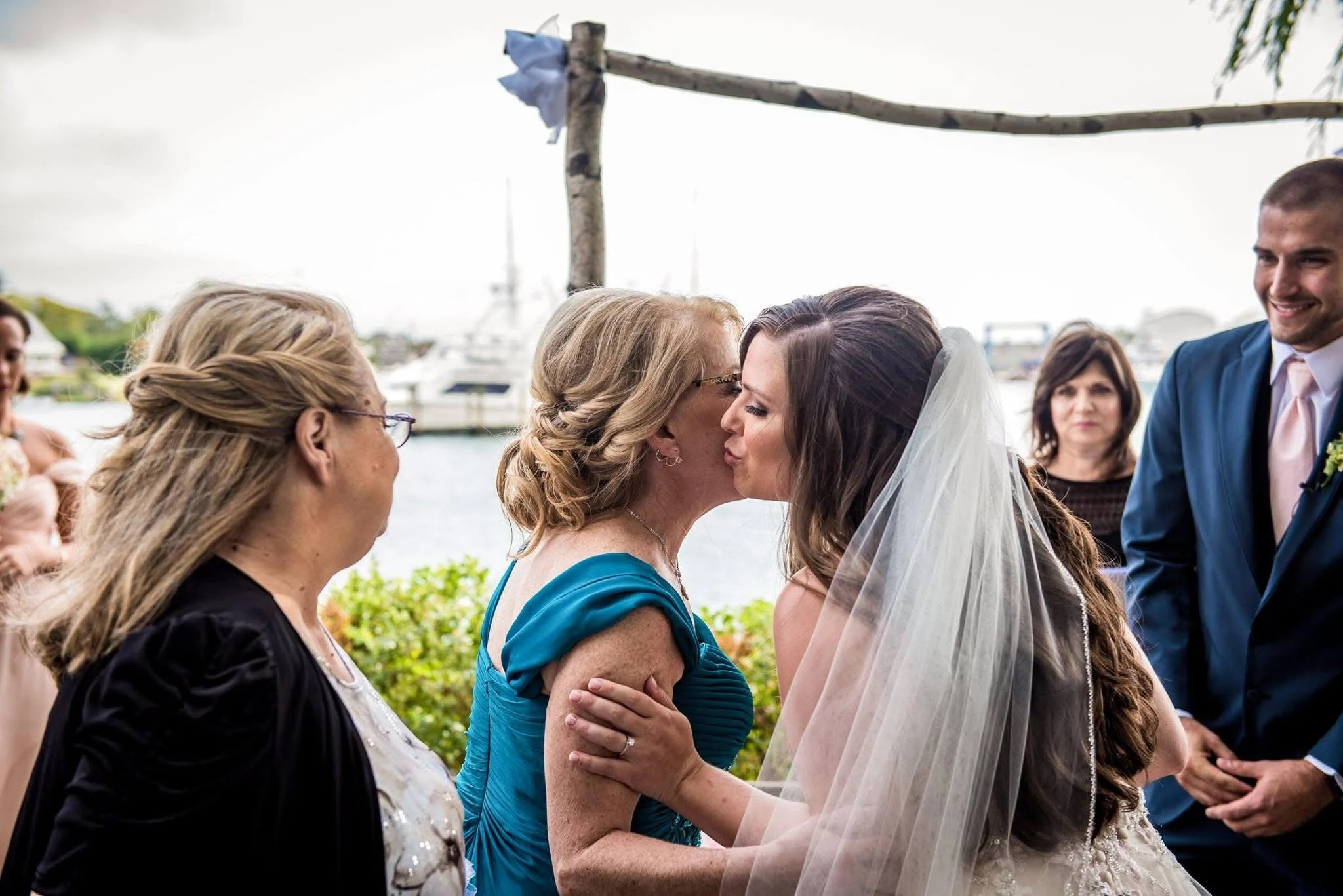 A bride and another woman sharing a kiss during a wedding ceremony outdoors near water, with guests in the background.