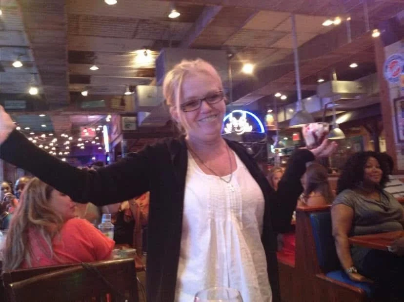 A woman smiling and holding ice cream in a restaurant or bar setting with people seated at tables in the background.