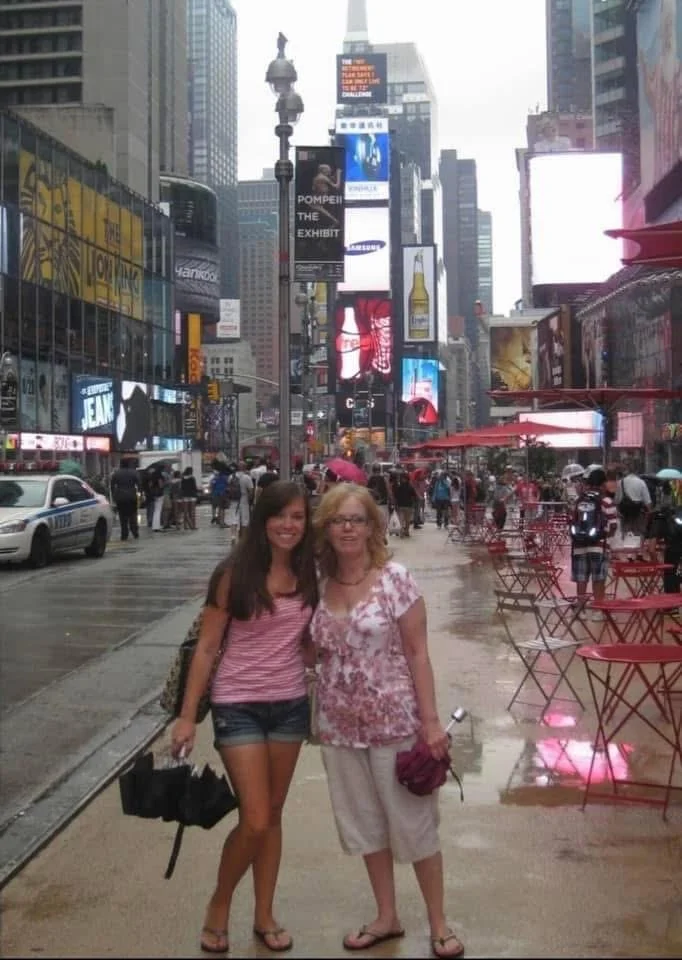 Two women standing on a city sidewalk in Times Square, New York City, with bright digital billboards and crowds in the background on a rainy day.