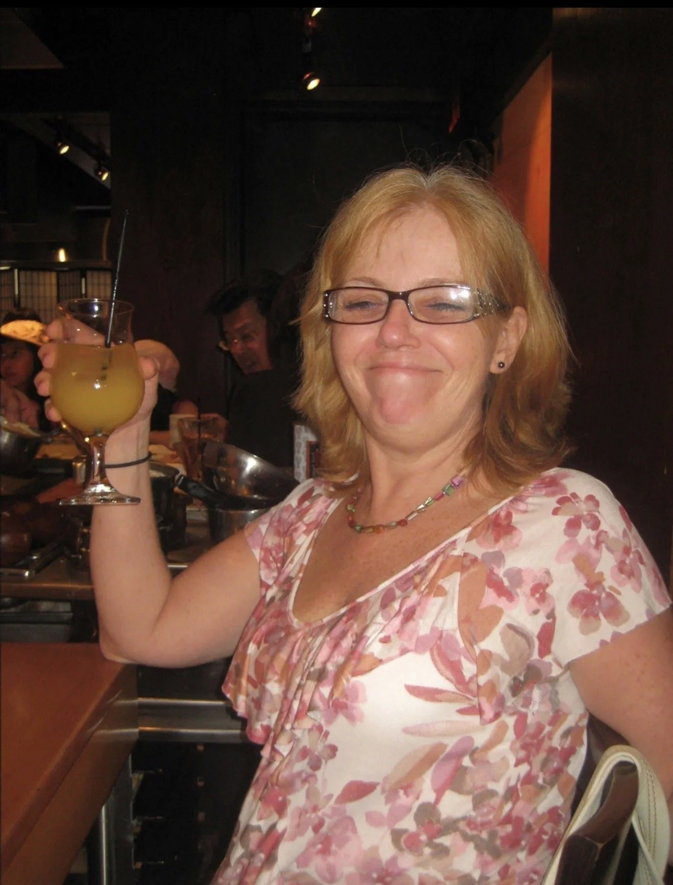 A woman with short red hair and glasses smiling while holding a yellow drink with a straw at a social gathering.